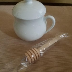 May include: White ceramic honey pot with a lid and a wooden honey dipper wrapped in plastic.