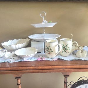 May include: A collection of white ceramic tableware and decorative items, including a two-tiered serving tray, bowls, mugs with green floral designs, and swan figurines. The items are arranged on a wooden table with a floral patterned runner.