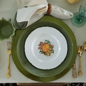 May include: A green and white dinner plate setting with a white plate featuring a fruit design, a green charger plate, and a white napkin with a brown napkin ring.