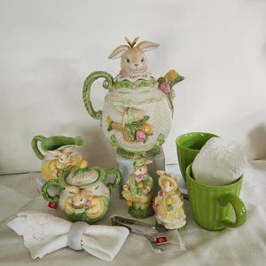 May include: A tea set featuring a teapot, sugar bowl, creamer, and two mugs. The teapot is shaped like a bunny and is decorated with floral and pastel colors. The sugar bowl and creamer are also bunny-shaped and feature similar floral designs. The mugs are green and feature a white bunny design. The set is perfect for a spring or Easter tea party.