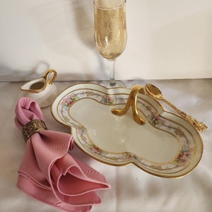 May include: A collection of elegant serving pieces. Includes a cream-colored, floral-decorated serving dish with gold trim, a gold-handled spoon, a pink napkin in a decorative ring, a small creamer, and a champagne flute with etched design.