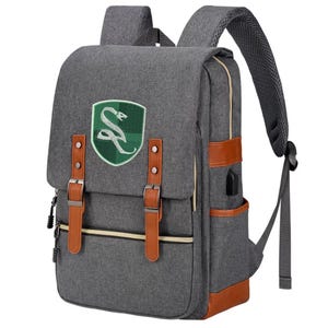May include: A grey backpack featuring a green shield-shaped patch with a white snake emblem. The bag includes brown faux leather straps with metal buckles, a front zippered pocket, side pockets, and padded shoulder straps with a mesh lining.