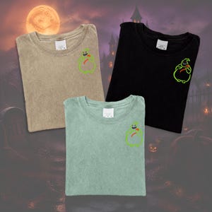 Könnte beinhalten: Drei T-Shirts mit Rundhalsausschnitt in Schwarz, Hellgrün und Beige, jeweils mit einer kleinen gestickten grünen Figur mit rotem Akzent auf der linken Brust. Die T-Shirts haben kurze Ärmel und einen Standardkragen und sind flach ausgelegt.