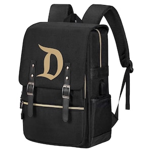 May include: Black backpack with a gold letter "D" on the front flap. The backpack has adjustable shoulder straps, a zippered front pocket, and two buckle closures. It features a USB charging port and a side pocket.