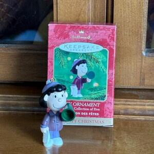 May include: A Hallmark Keepsake ornament featuring a cartoon character in a purple coat, blue pants, and a green megaphone. The ornament is next to its original box, which has a picture of the character and the words "Keepsake Ornament".