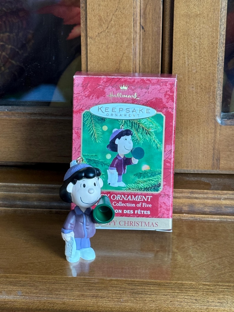 May include: A Hallmark Keepsake ornament featuring a cartoon character in a purple coat, blue pants, and a green megaphone. The ornament is next to its original box, which has a picture of the character and the words "Keepsake Ornament".