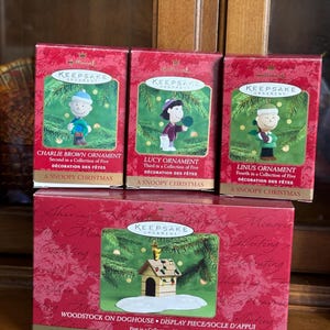 May include: Four Hallmark Keepsake Christmas ornaments featuring Peanuts characters. The boxes are red with gold lettering and images of Charlie Brown, Lucy, Linus, and Woodstock on a doghouse. Each box is labeled "A Snoopy Christmas."