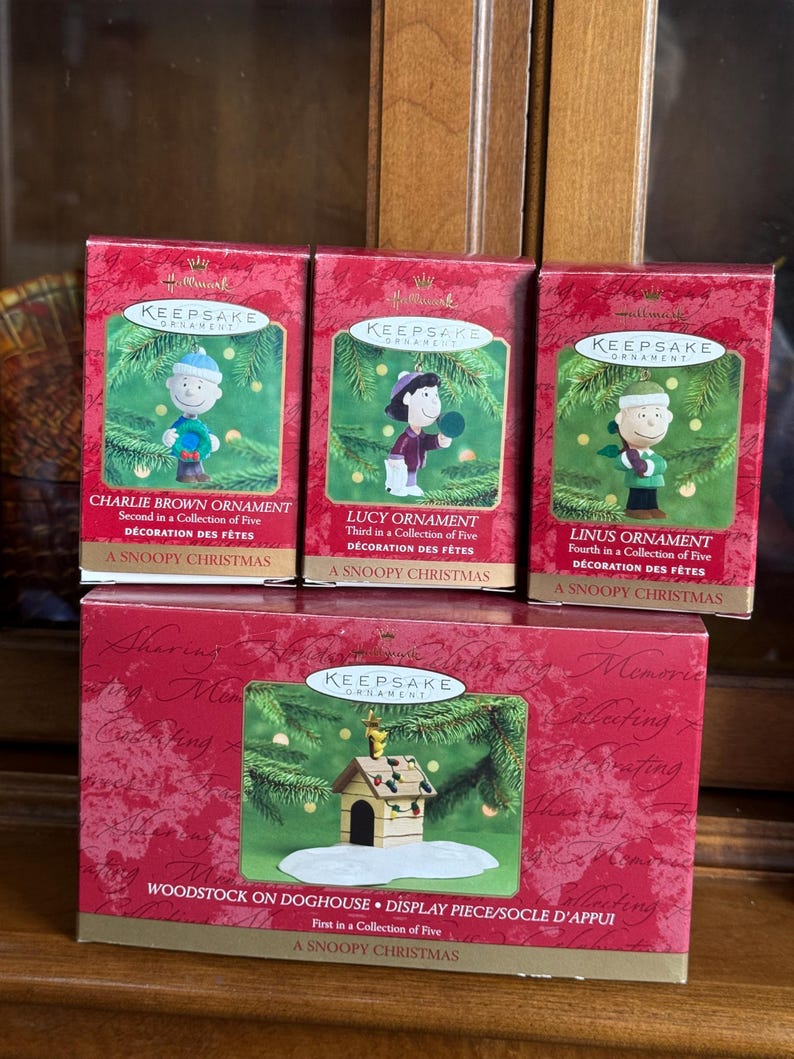 May include: Four Hallmark Keepsake Christmas ornaments featuring Peanuts characters. The boxes are red with gold lettering and images of Charlie Brown, Lucy, Linus, and Woodstock on a doghouse. Each box is labeled "A Snoopy Christmas."