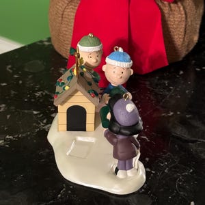 May include: A Christmas ornament featuring Charlie Brown, Snoopy's doghouse, and other Peanuts characters. The ornament has a snow-covered base and is decorated with lights. The characters are wearing winter hats and coats. A red bow is in the background.