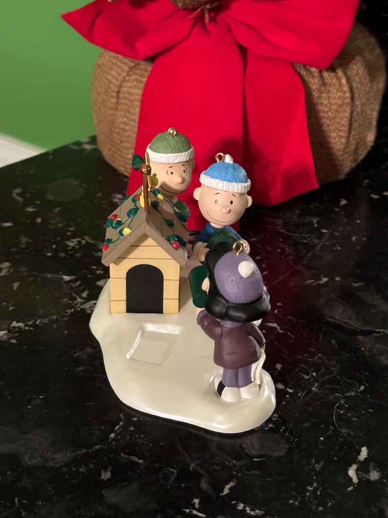 May include: A Christmas ornament featuring Charlie Brown, Snoopy's doghouse, and other Peanuts characters. The ornament has a snow-covered base and is decorated with lights. The characters are wearing winter hats and coats. A red bow is in the background.
