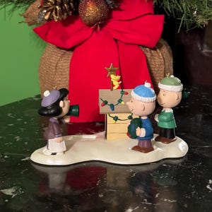 May include: A decorative Christmas ornament featuring Peanuts characters. Lucy holds a megaphone, while Charlie Brown and Linus decorate Snoopy's doghouse with lights. The ornament has a star with the year 2000 on top.