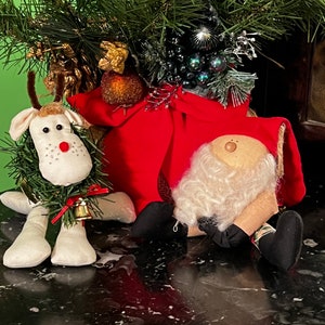 May include: Christmas decorations featuring a reindeer and Santa Claus plush dolls. The reindeer has a green wreath around its head and a small bell. Santa Claus has a white beard and is wearing a red suit. The background includes a green garland.