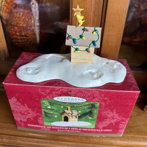 May include: A Hallmark Keepsake ornament featuring a wooden doghouse with a small yellow bird figure and a star that reads "2000". The doghouse is decorated with colorful Christmas lights and sits on a white cloud-shaped base. The box reads "Woodstock on Doghouse".
