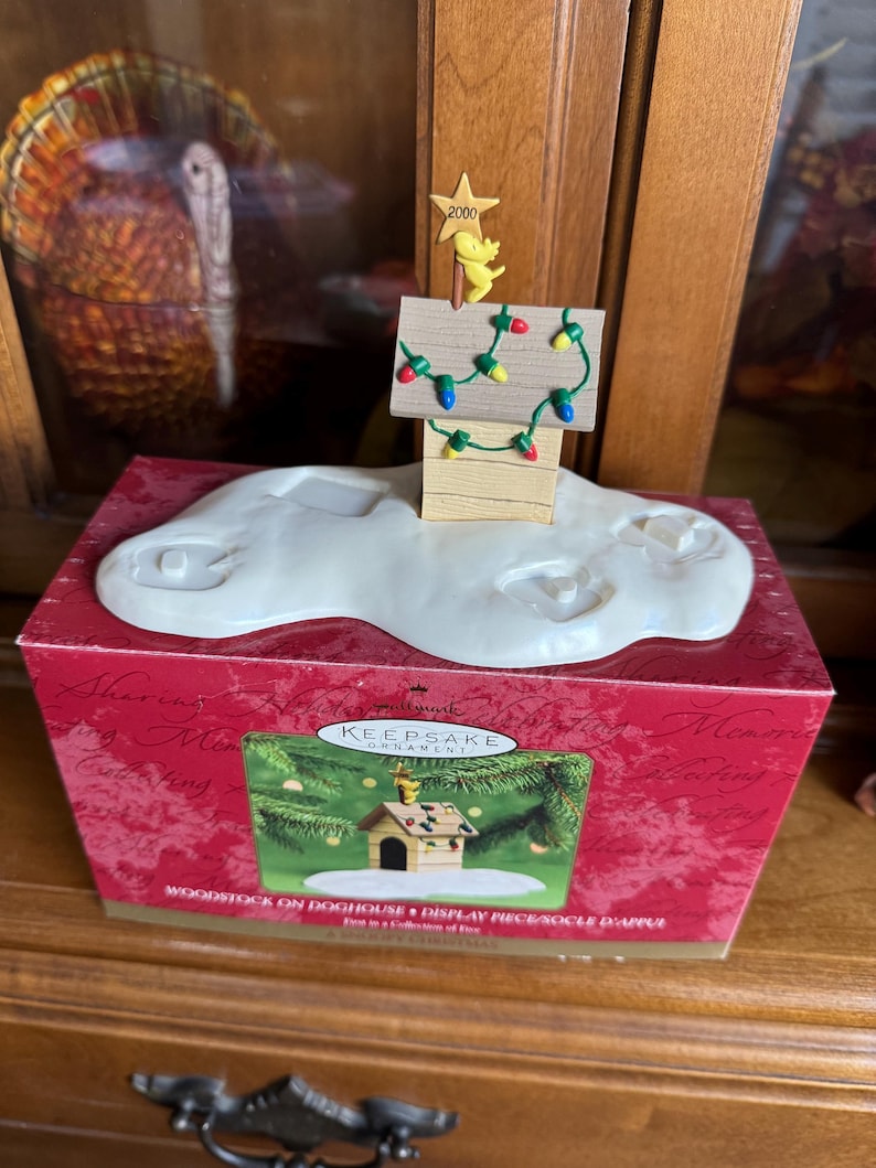 May include: A Hallmark Keepsake ornament featuring a wooden doghouse with a small yellow bird figure and a star that reads "2000". The doghouse is decorated with colorful Christmas lights and sits on a white cloud-shaped base. The box reads "Woodstock on Doghouse".