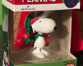 May include: A Snoopy Christmas tree ornament in a box. The white Snoopy figure wears a red hat and scarf with green glitter accents. The box reads "PEANUTS" and "Christmas Tree Ornament."