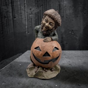 May include: A brown and grey figurine of a bearded character wearing an acorn-shaped hat, leaning over a carved orange pumpkin with a wide, dark, triangular-eyed grin. The base is textured and features the embossed text ROMPUNKIN on the front.