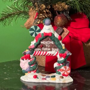 以下が含まれることがあります： 「WELCOME TO PLUM PUDDIN PLACE」の文字が入った、家の形をしたセラミック製のクリスマスオーナメント。家はキャンディー、リボン、赤と白のストライプの日よけで飾られています。デザイン全体に緑と赤のアクセントが使用されています。