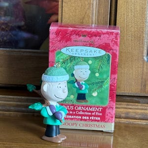 May include: A Hallmark Keepsake ornament featuring a Charlie Brown figure in a green outfit, holding a branch with purple berries. The ornament is next to a red box with the words "Keepsake Ornament" and "Snoopy Christmas."