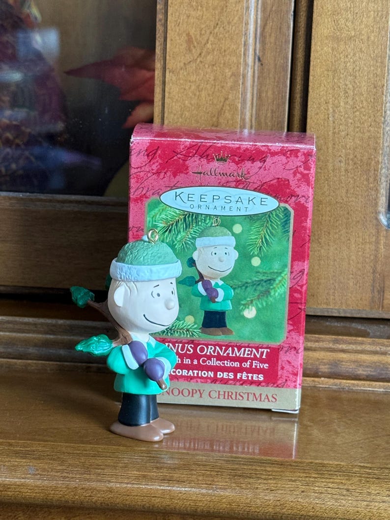 May include: A Hallmark Keepsake ornament featuring a Charlie Brown figure in a green outfit, holding a branch with purple berries. The ornament is next to a red box with the words "Keepsake Ornament" and "Snoopy Christmas."