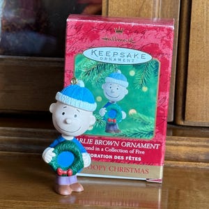 May include: A Charlie Brown Christmas ornament figurine, wearing a blue hat and holding a green wreath with a red bow. The ornament is in front of a red Hallmark Keepsake box.