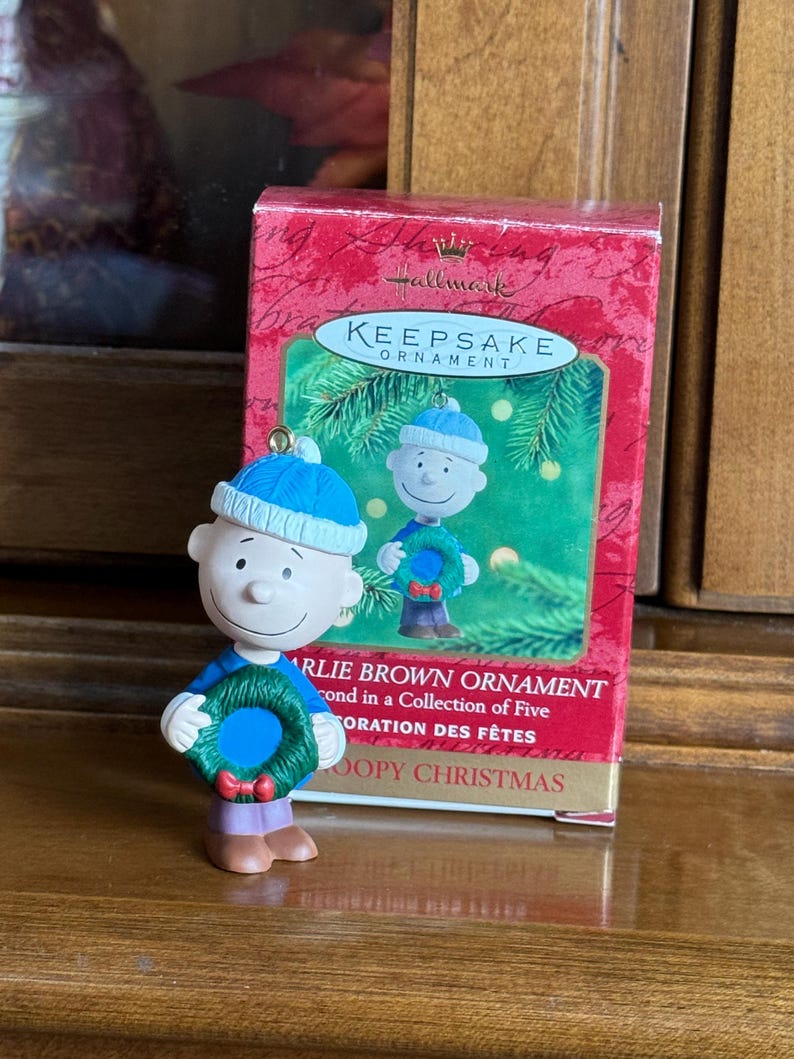 May include: A Charlie Brown Christmas ornament figurine, wearing a blue hat and holding a green wreath with a red bow. The ornament is in front of a red Hallmark Keepsake box.