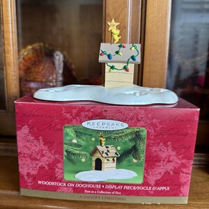 May include: A Snoopy Christmas display piece featuring Woodstock on a doghouse, adorned with colorful lights and a star. The ornament sits atop a snow-covered base, presented in a red box with the text "WOODSTOCK ON DOGHOUSE".