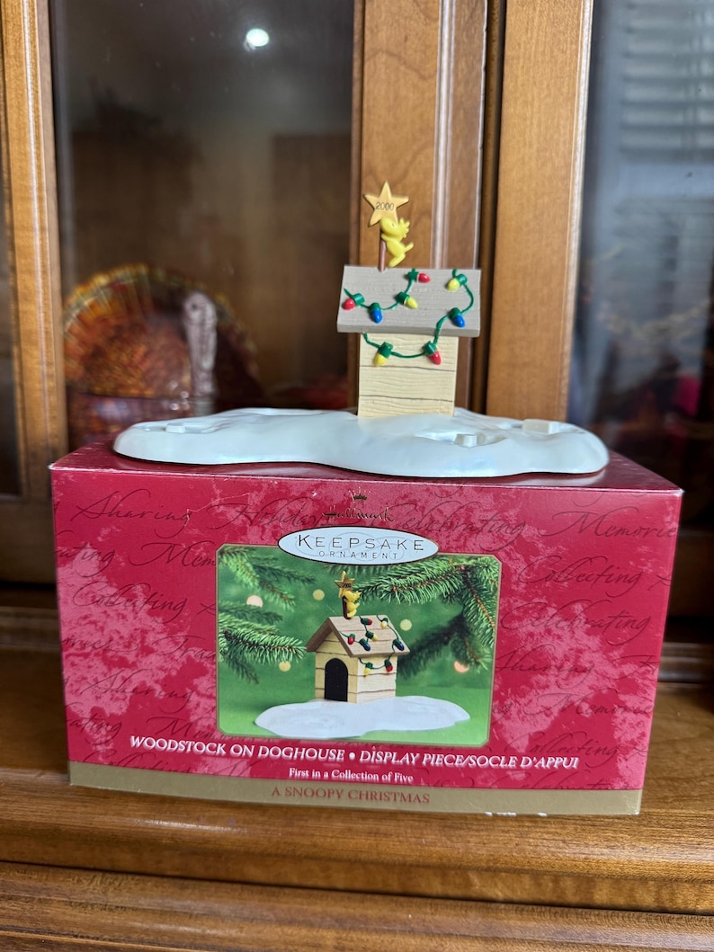 May include: A Snoopy Christmas display piece featuring Woodstock on a doghouse, adorned with colorful lights and a star. The ornament sits atop a snow-covered base, presented in a red box with the text "WOODSTOCK ON DOGHOUSE".