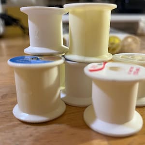 May include: A collection of off-white and pale yellow thread spools. Some spools have labels with text in red and blue. The spools are stacked on a wooden surface, suggesting a sewing or craft theme.