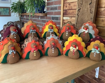 Puede incluir: Una colección de adornos de pavo hechos a mano con fieltro. Cada pavo tiene un cuerpo marrón, una cola colorida y una pajarita. Las colas son en tonos naranja, amarillo y rojo. Los pavos están colocados sobre una superficie de madera.