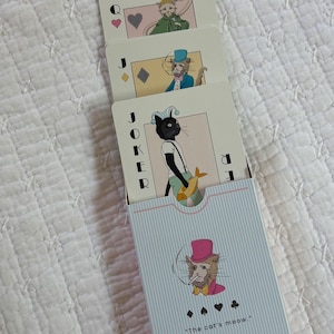 May include: A deck of playing cards featuring whimsical illustrations of cats dressed in royal attire. The cards are partially out of a light blue striped box with the text "The cat's meow."