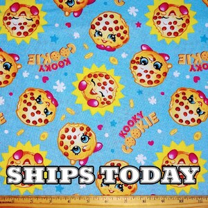 May include: A blue fabric with a repeating pattern of smiling cookie characters. The cookies are yellow with red and white sprinkles. The text "Kooky Cookie" is repeated throughout the fabric.