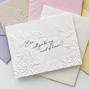 Peut inclure: Une carte blanche avec des motifs floraux en relief et le texte noir Erin, will you be my maid of honour? entourée de plusieurs enveloppes aux tons pastel lavande, beige, rose, crème et jaune, chacune avec des motifs floraux en relief.