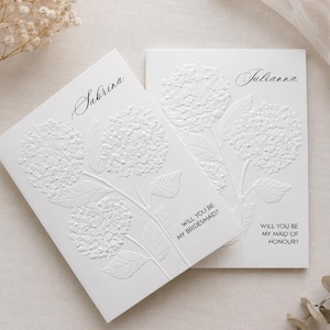 Op de afbeelding: Twee witte rechthoekige kaarten met in reliëf gedrukte hortensia bloemmotieven. Eén kaart is gepersonaliseerd met de naam Sabrina en de tekst WILL YOU BE MY BRIDESMAID? De andere kaart bevat de naam Julianna en WILL YOU BE MY MAID OF HONOUR?