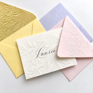 May include: A white rectangular card with the name Lauren printed in black script, featuring an embossed floral pattern. The card is surrounded by four envelopes in gold, pale yellow, light purple, and soft pink, each with an embossed floral design.