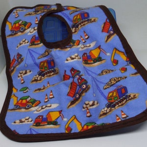 May include: A blue and brown bib with a construction theme. The bib features a variety of construction vehicles, including trucks, excavators, and cranes. The bib has a hole in the center for the child's head.