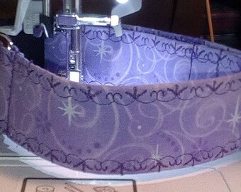 May include: A close-up of a purple fabric being sewn on a sewing machine. The fabric has a white swirl pattern and a purple zig-zag stitch around the edge.