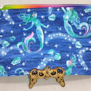 May include: A blue zippered pouch featuring a mermaid design with a rainbow zipper. The pouch has a blue background with white stars, seashells, and mermaids in shades of teal, pink, and purple.