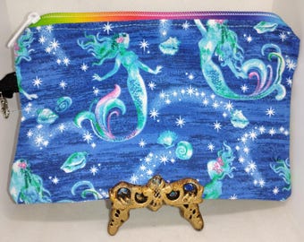 May include: A blue zippered pouch featuring a mermaid design with a rainbow zipper. The pouch has a blue background with white stars, seashells, and mermaids in shades of teal, pink, and purple.