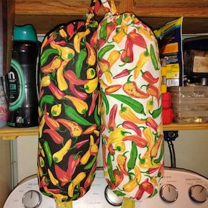 Op de afbeelding: Twee stoffen groentezakken met trekkoord, hangend aan een witte knop. Eén tas heeft een zwarte achtergrond met kleurrijke chilipepers. De andere tas heeft een witte achtergrond met chilipepers.