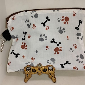 May include: White zippered pouch with a brown zipper and a black strap with a silver clip. The pouch has a white background with a repeating pattern of black bones and gray and brown paw prints.