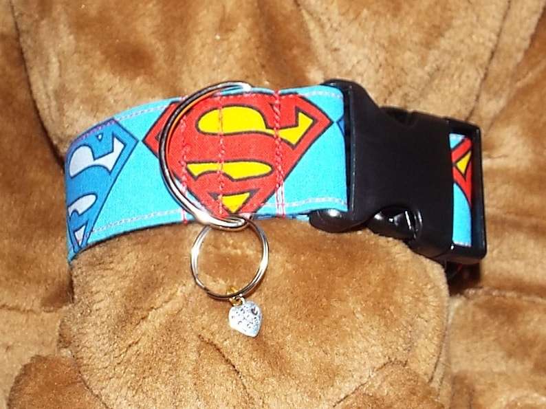 May include: A blue and red dog collar with a Superman logo and a silver ring with a heart charm attached.
