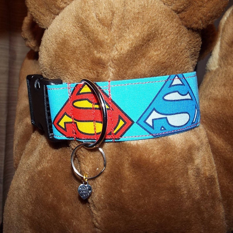 May include: A blue dog collar with red, yellow, and blue Superman logos. The collar has a silver D-ring and a silver heart charm attached.