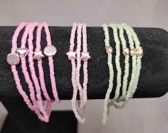 Könnte beinhalten: Drei übereinanderliegende Armbänder mit unterschiedlich farbigen Perlen und Charms. Das erste Armband ist rosa mit silbernen Herz- und Stern-Anhängern. Das zweite Armband ist grün mit silbernen Stern-Anhängern. Das dritte Armband ist grün mit goldenen Anhängern.
