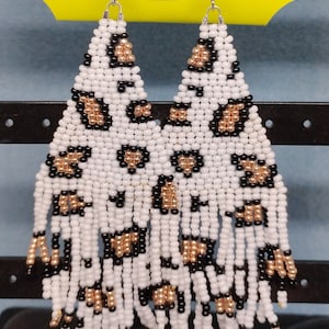 May include: A pair of white beaded earrings with a leopard print pattern. The earrings are made with white, black, and gold beads and have a fringe design.