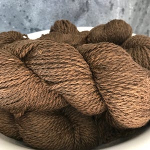 May include: A close-up of a brown yarn skein in a white bowl. The yarn is soft and fluffy, and it is perfect for knitting or crocheting.