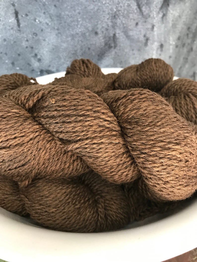 May include: A close-up of a brown yarn skein in a white bowl. The yarn is soft and fluffy, and it is perfect for knitting or crocheting.