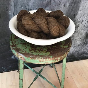 May include: A white ceramic bowl filled with brown yarn sits on a green metal stool. The stool has a distressed finish and is made of metal.