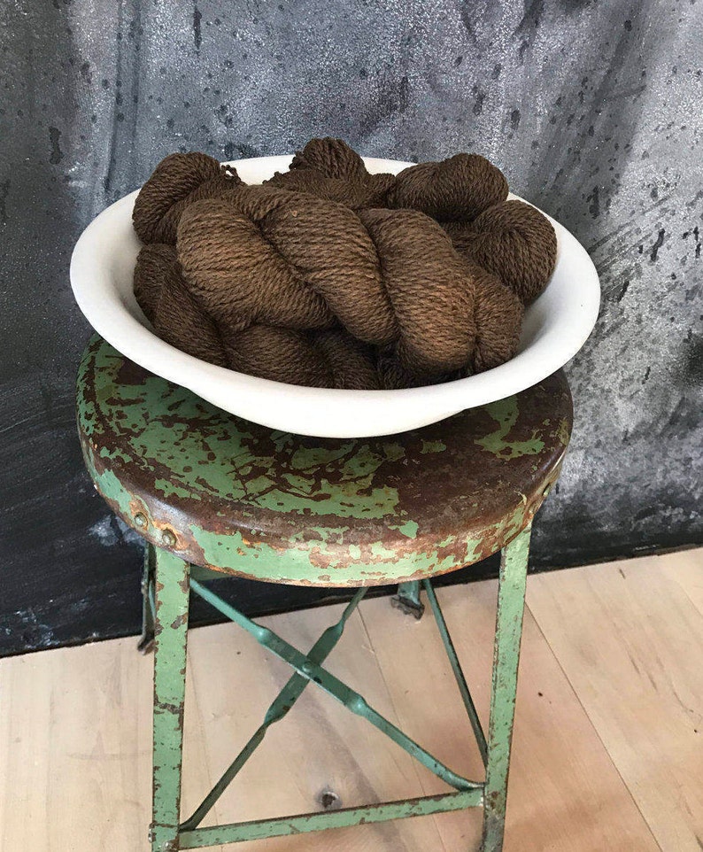 May include: A white ceramic bowl filled with brown yarn sits on a green metal stool. The stool has a distressed finish and is made of metal.