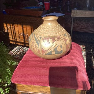 May include: A brown ceramic vase with intricate geometric designs in black, white, and red. The vase is sitting on a red velvet cushion.