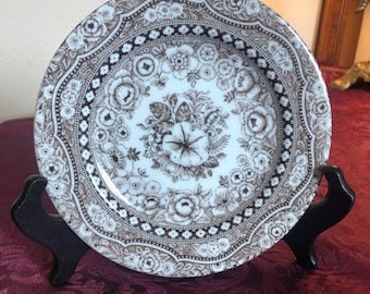 May include: A white ceramic plate with a brown floral pattern. The plate has a scalloped edge and is decorated with a repeating pattern of flowers and leaves.